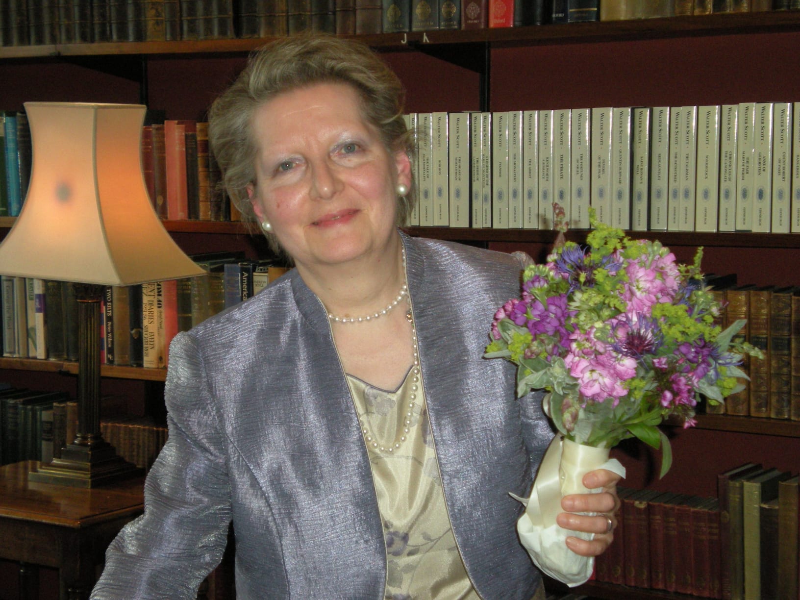 This is a photograph of Laura Scott, she is wearing a cream dress and purple blazer. She is picture in front of a bookshelf and a lamp and is holding a bunch of flowers that are pink and purple.