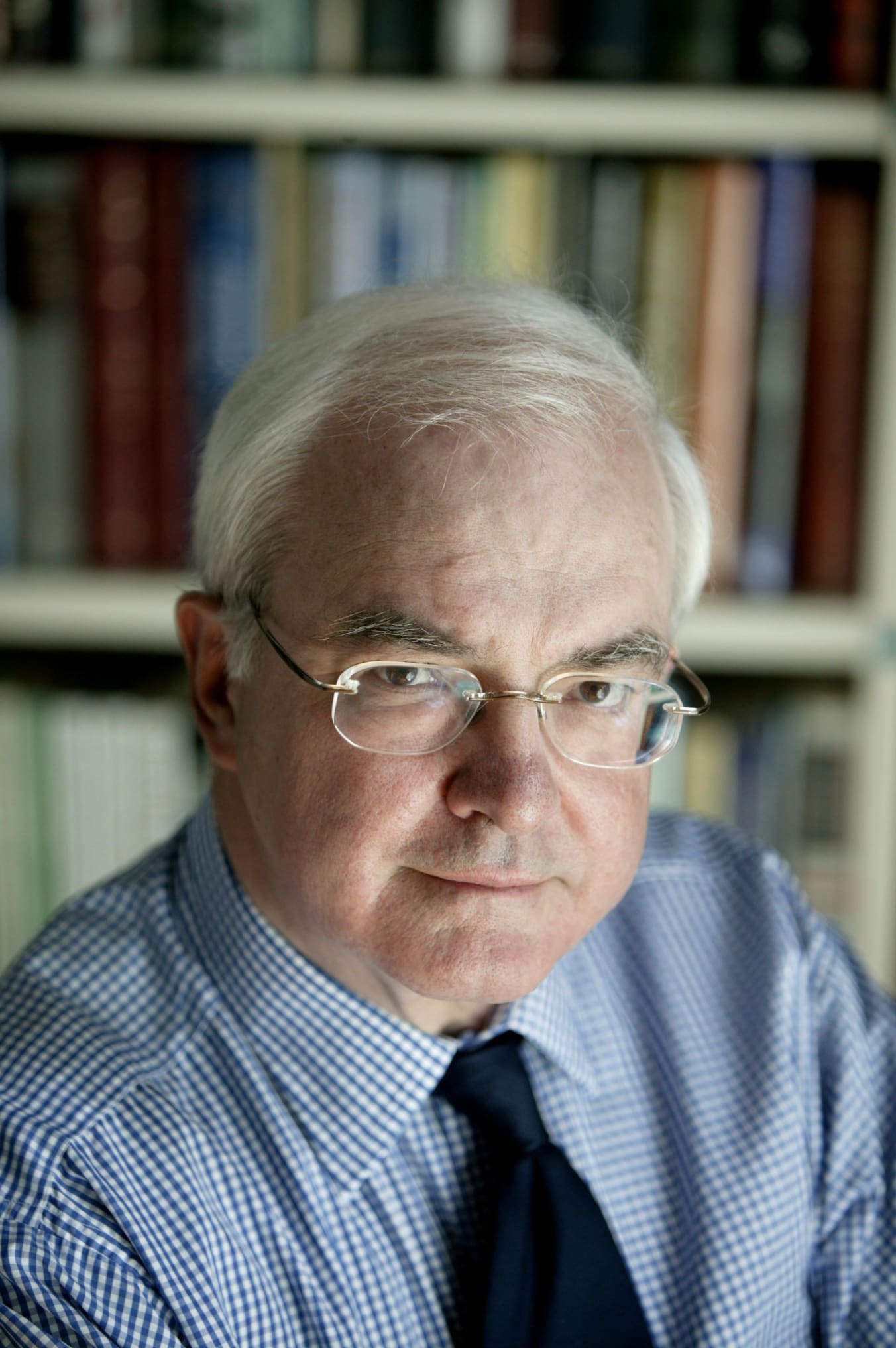 This is a photograph of Magnus Linklater. He is wearing glasses, a blue shirt and navy tie. He is pictured seated with a bookcase behind him.