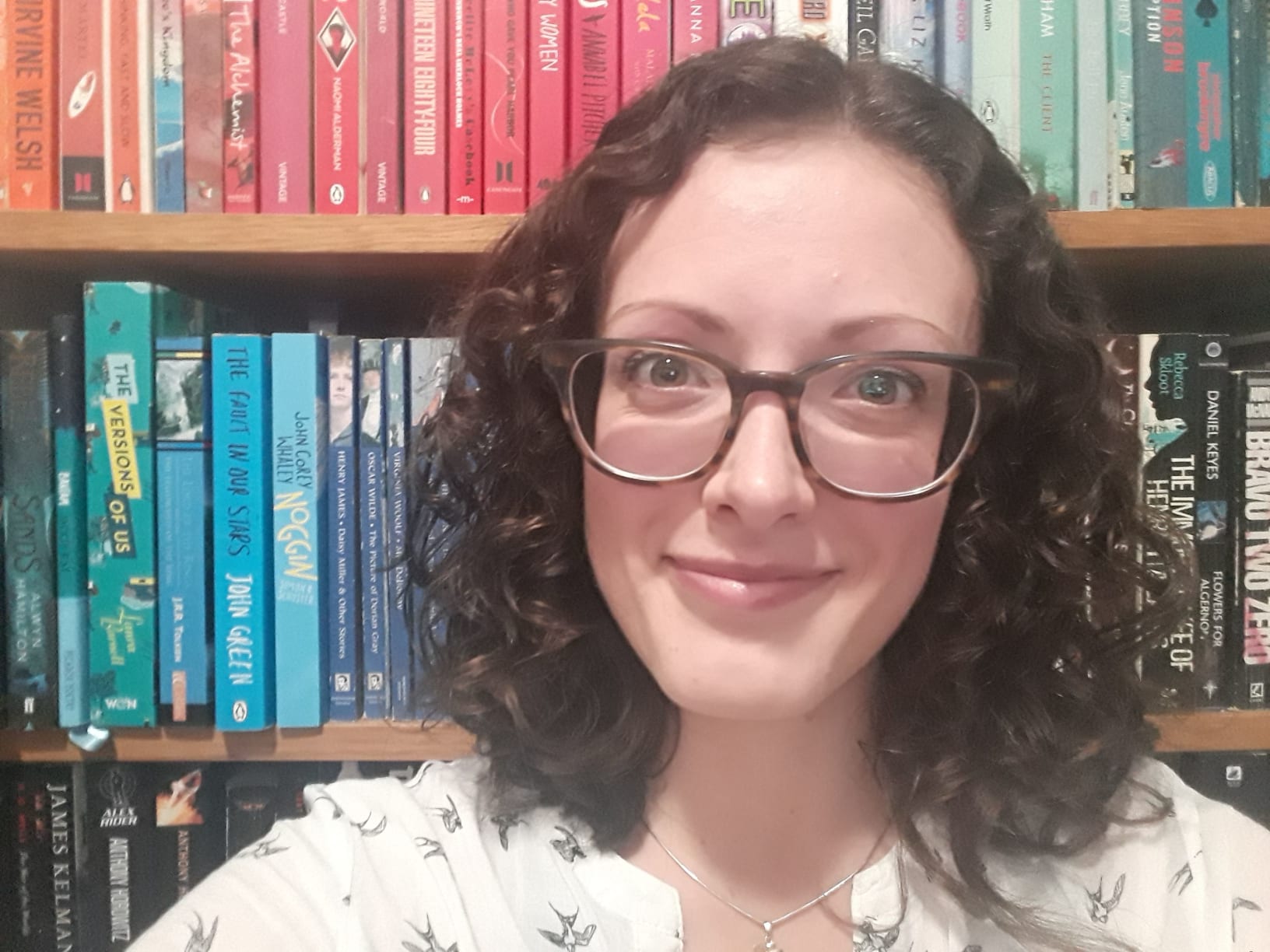 This is a photograph of Kirstin Lamb who has shoulder-length, brown, curly hair and is wearing glasses. She is pictured in from of a bookshelf with brightly coloured books on it.